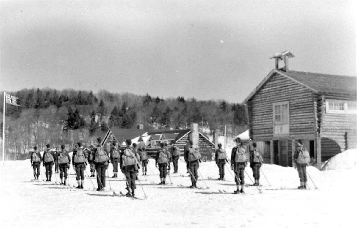 En tropp med rekrutter som står på en plass foran noen tømmerhytter. Rekruttene har ski bå beina og ryggsekk på seg..Snø på bakken.