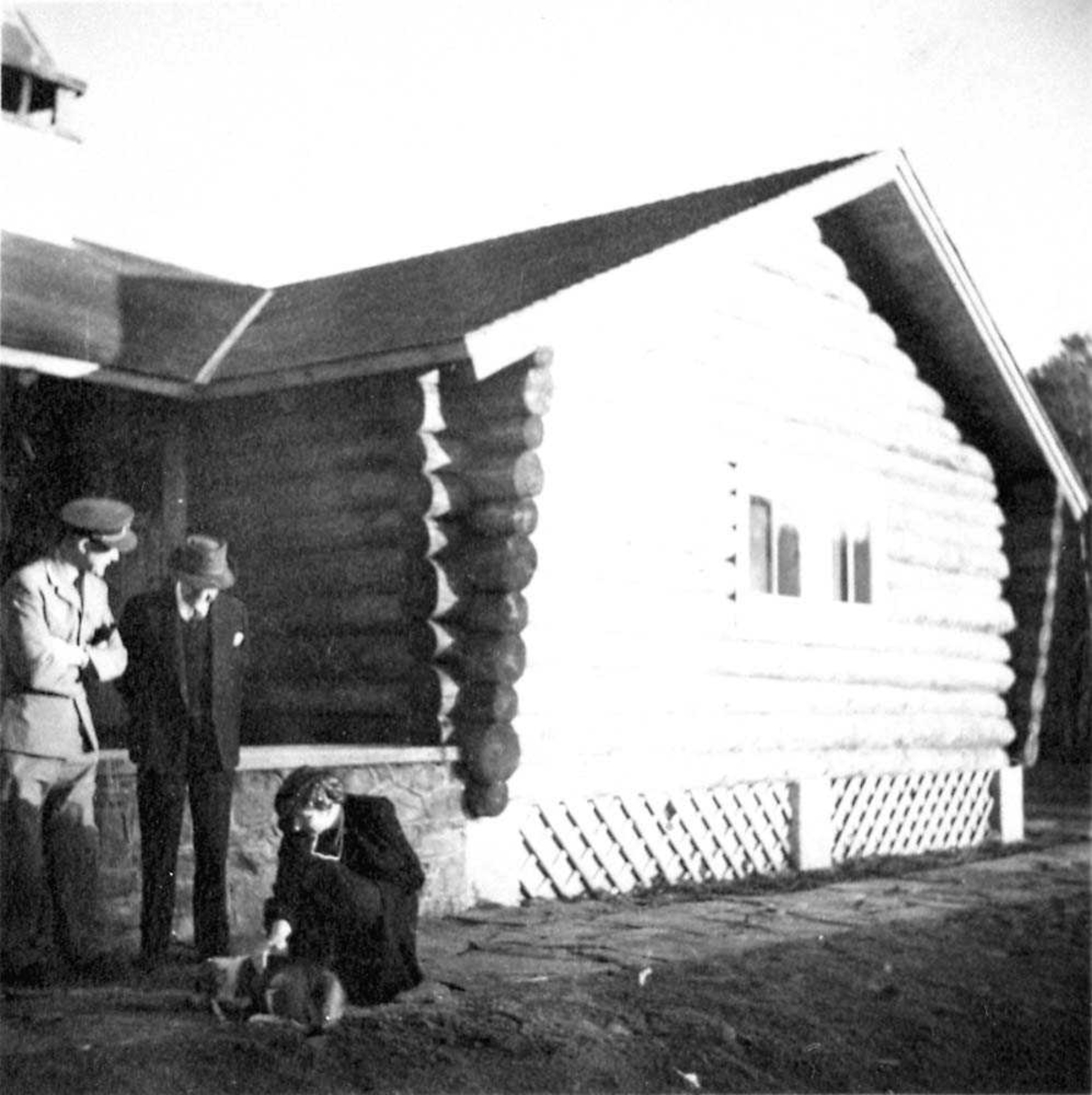 Portrett. Tre personer, to menn og en kvinne foran en bygning. Kvinnen klapper en hund.