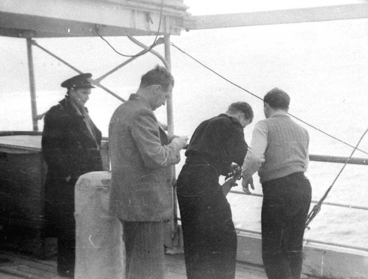 Fire personer som står på dekket til en båt, menn. En av mennene har et skytevåpen.