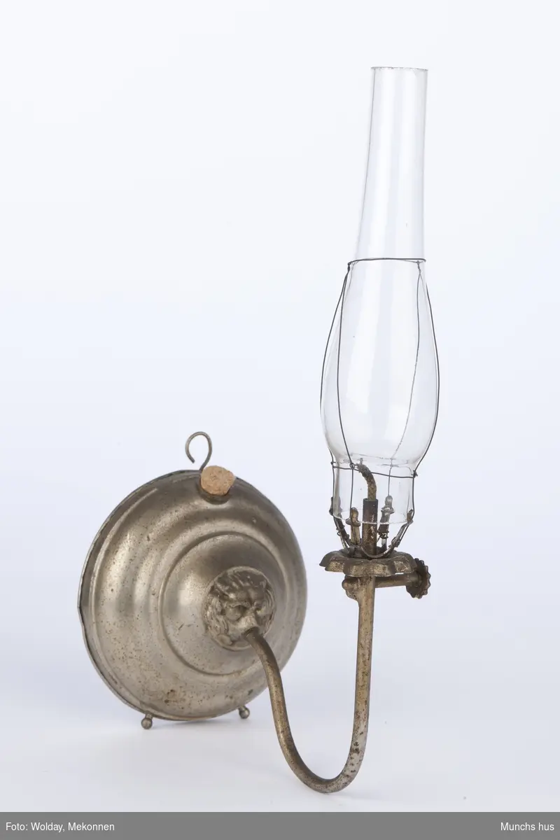 Parafinlampe. Opprinnelig glasskuppel knust, men skiftet ut med nytt på senere tidspunkt. Lampen kan stå eller henge på vegg.
Kuppelholder er festet i et løvehode på beholder.