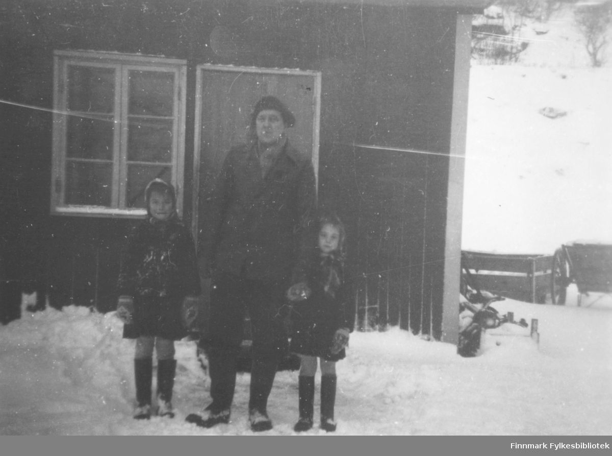 Evald Valen og hans to døtre?, står foran butikkbrakka i Lillefjord. Det er vinter og snøen ligger på bakken. De er godt kledt. Til høyre for brakka, står en kjerre