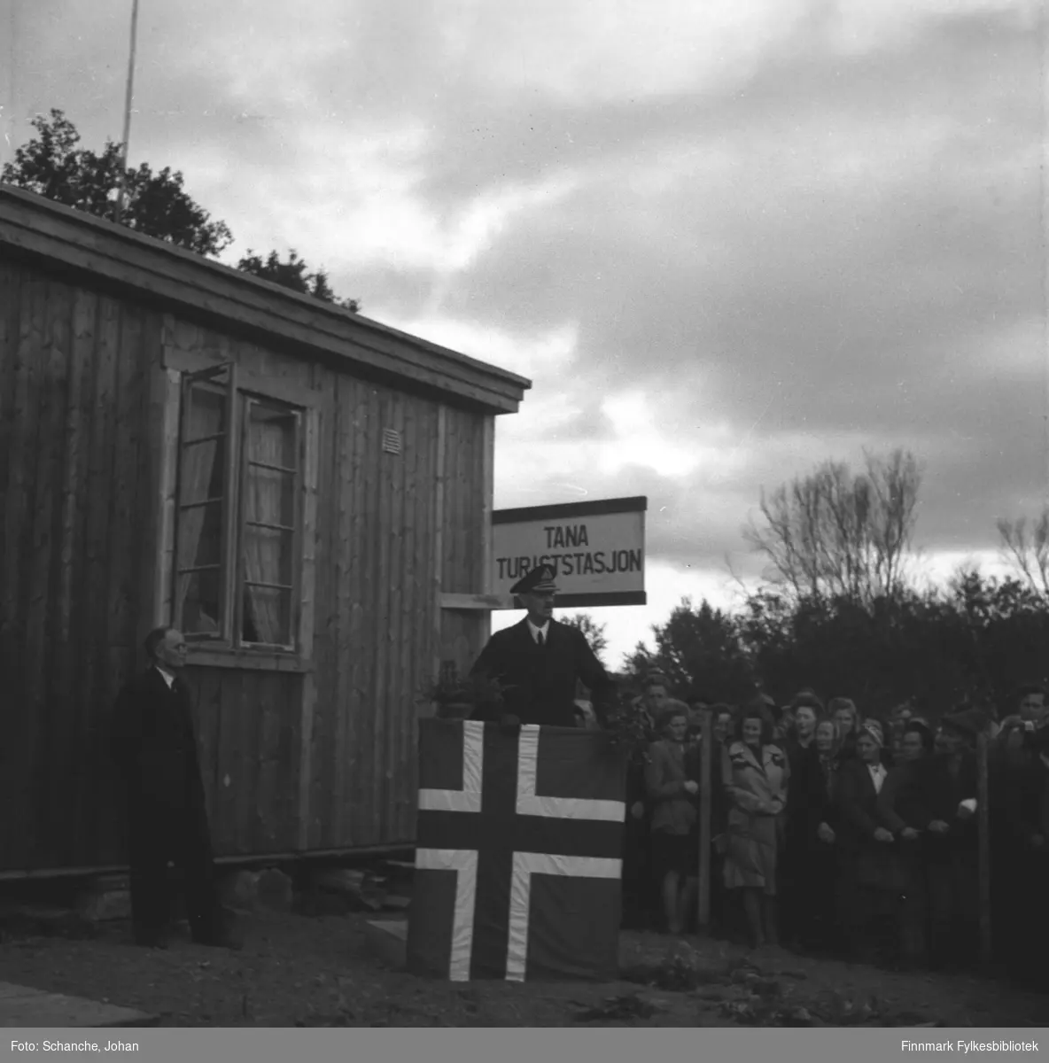 Kongebesøk i 1946:  Kong Haakon VII besøker Tana. Kongen står på talerplattformen ved Tana Turiststasjon og holder tale til folk som har samlet seg bak en gjerde like ved. Talerstolen er dekorert med flagg. Selve turiststasjonen er en nybygdt brakke.