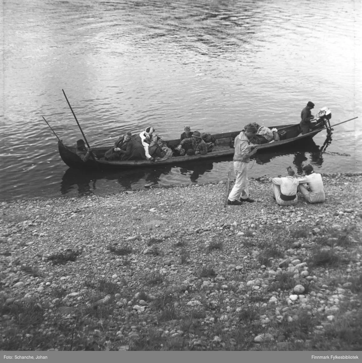 En gruppe mennesker i turklær sitter på elvebåten som er i ferd med og avgå. På båten ryggsekker og bagasje. To menn sitter  og en stårpå elvekanten. Bildet er tatt fra elvebredd ned mot båten.