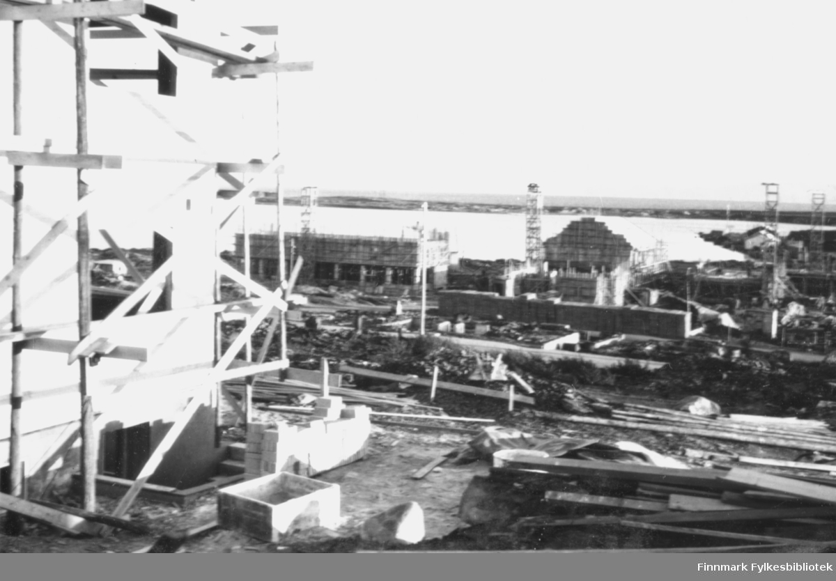 Gjenoppbygging av Vadsø sentrum etter andre verdenskrig. I forgrunnen til venstre i bildet står et hus under bygging med diverse byggematerialer rundt. I bakgrunnen ser man flere hus under oppbygging med stillaser rundt, og forskallinger til bygging av piper står flere steder i området. Utsikt mot havet og Vadsøya