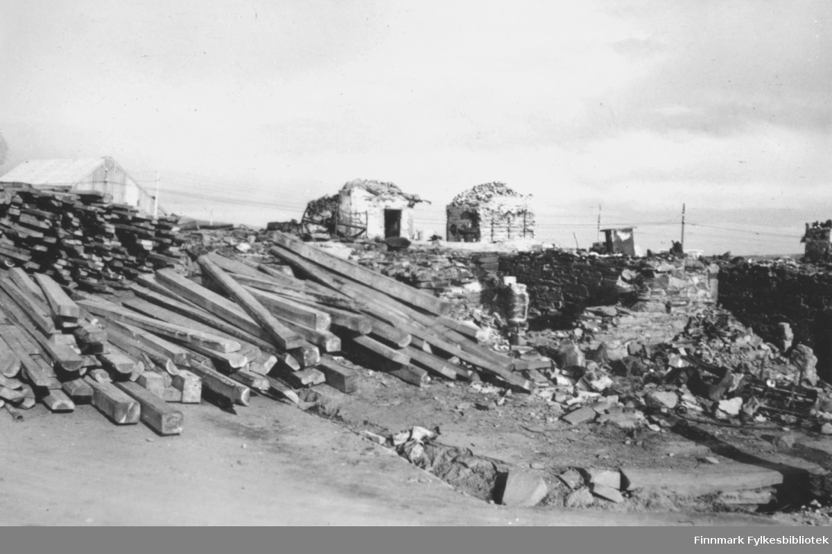 Fra Vadsø sentrum høsten 1946. Materialer har blitt fraktet inn for å starte gjenreisningen av alle bygningene som ble bombet under andre verdenskrig. I forgrunnen ligger hauger med treplanker oppå restene av en grunnmur. I bakgrunnen er det flere ruiner, en brakke og noen stolper