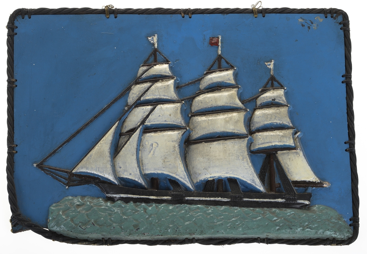 Rektangulært bilde av ei seilskute laget av støpejern. Motivet er deretter malt. Rundt bildet er det festet på tvinnet og behandlet tau.