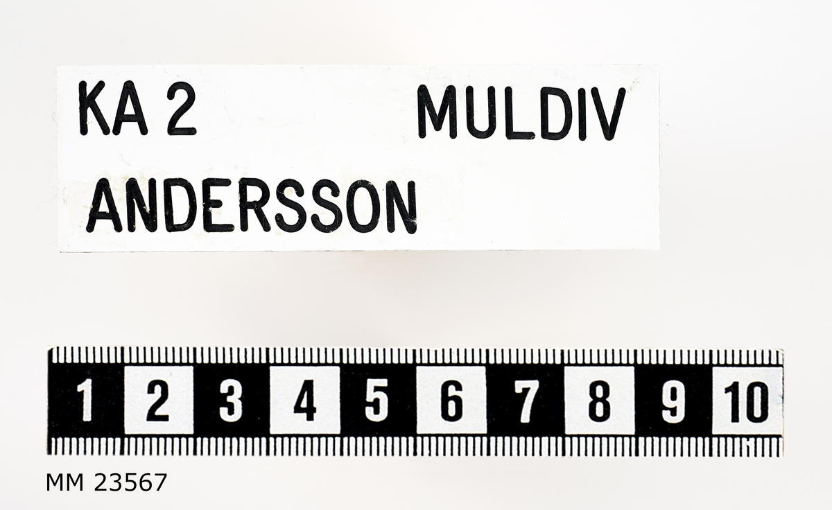 Namnbricka i plast med vit bakgrund och svart text. På brickan står på översta raden "KA 2 MULDIV" och på andra raden "ANDERSSON". Texten är graverad. Namnbrickan saknar bygel för fäste i uniform.