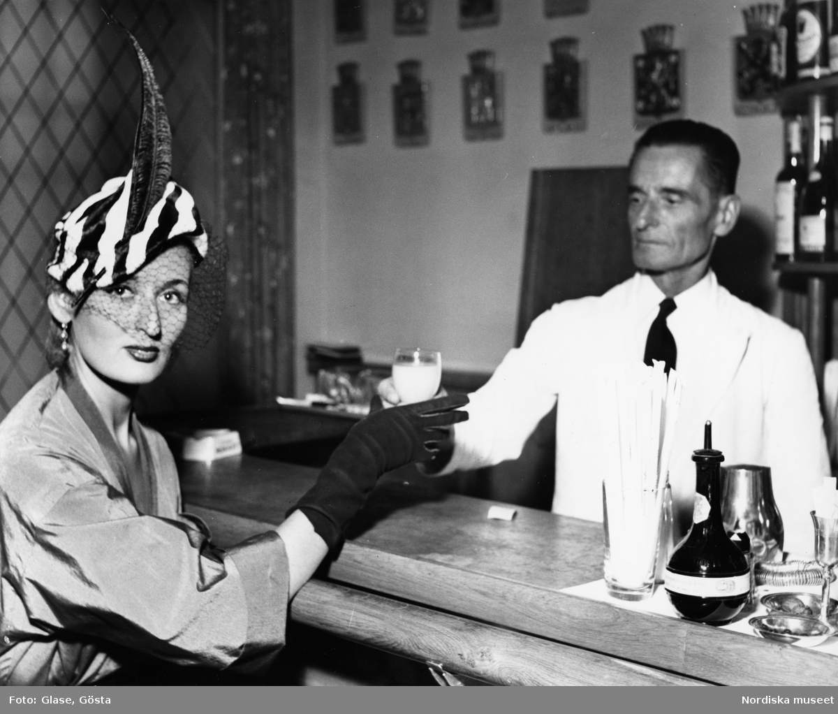 Dammode. Mannekäng, Kim Söderlund, blir serverad i en bar, klädd i zebramönstrad hatt med fjäder och flor och handskar. Man i vit kavaj.