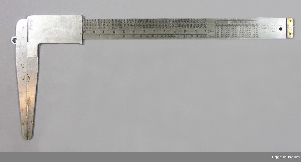 Taksasjonsklave i rutfritt stål. Linjal med flere forskjellige tabeller for gran og furu. 4 utregningstabeller. Messingkloss i enden av linjal.