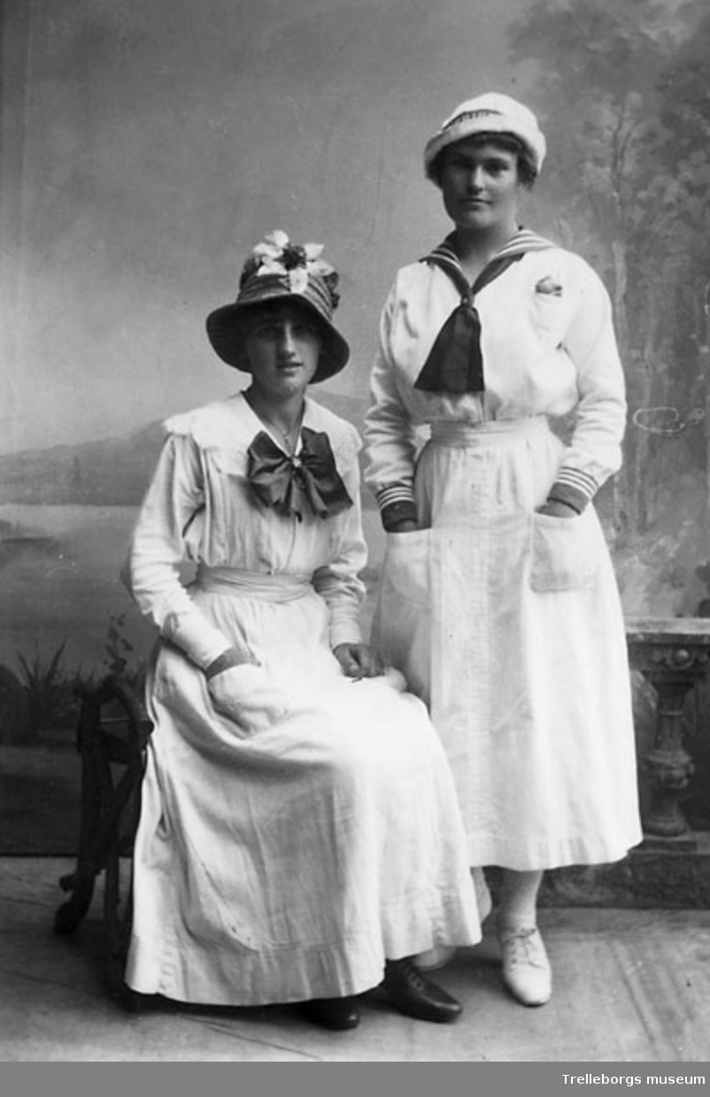 Två unga kvinnor i ljusa klänningar. En i hatt med smatl brätte och hög kulle prydd med en blomma, en i sjömansblus och mössa.