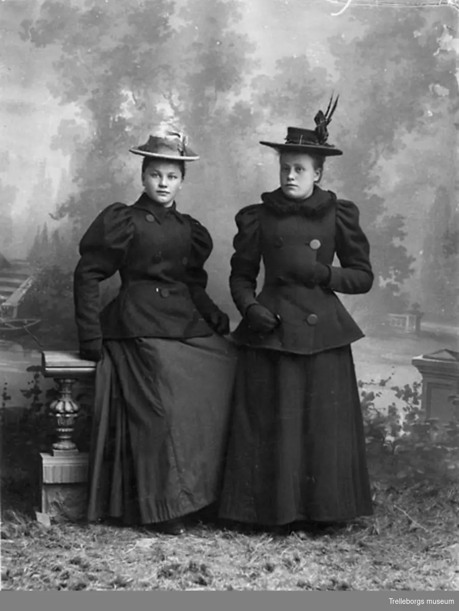 Två kvinnor i tvådelad dräkt med dubbelknäppt jacka, åtsittande ärmar med en lättare puff högst upp, handskar och hatt.