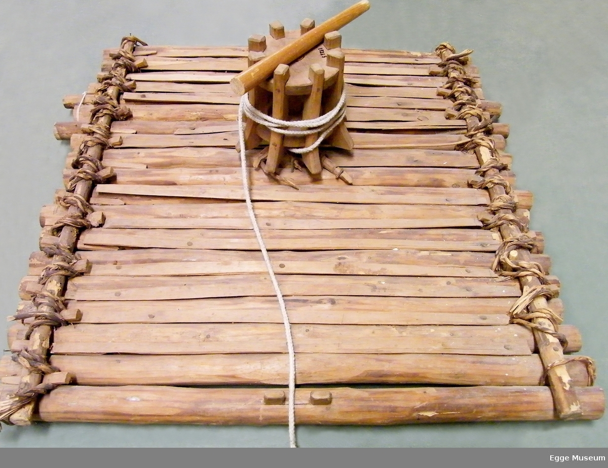 Modell av spillflåte laget i rundbarket gran. Flåten er bygd av rundtømmer som er kledd med halvklovninger. Tømmerstokkene er festet med vidjespenninger, som er kilt fast til stokker med store kiler stukket inn i løkker laget av vidjespenning. Ubehandlet. Flåten er tilnærmet kvadratisk i form. Midt på flåten står "jomfrua" eller tønna med oppkveilet tau. Det følger med en dreg laget av ståltråd. Ingen årer. Sammenføyningen med vidjebånd gjorde at flåten tålte bølger. Det er ukjent hvem som har laget modellen. Ukjent modellformat.