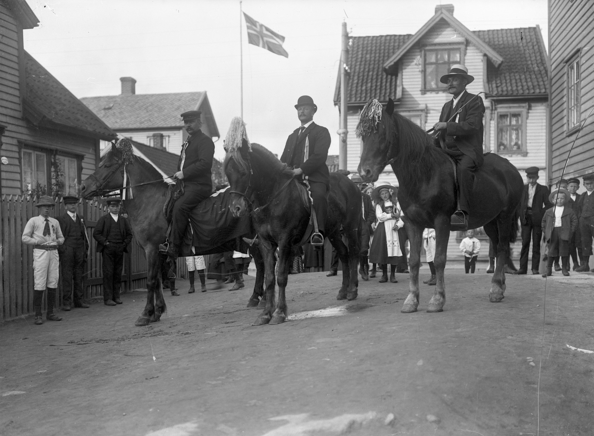 Tre ryttere på hest 17. mai. Det norske flagget vaier på en flaggstang midt på bildet. Hus i bakgrunnen. Barn og noen få voksne bak rytterne og til venstre.