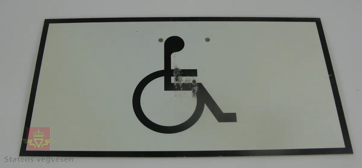 Rektangulært skilt av tre mm. aluminium. Svart bård og svart symbol, (rullestol), på hvit lysreflekterende bunn. Grå bakside. Skiltet har fire hull for fastskruing.