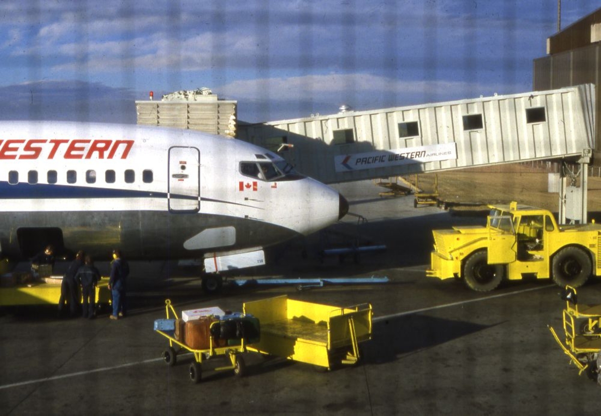 Lufthavn/Flyplass. Et fly fra Pasific Western, Canada,  parkert ved flyplassterminalen. Stor aktivitet ved flyet, inn eller utlasting.