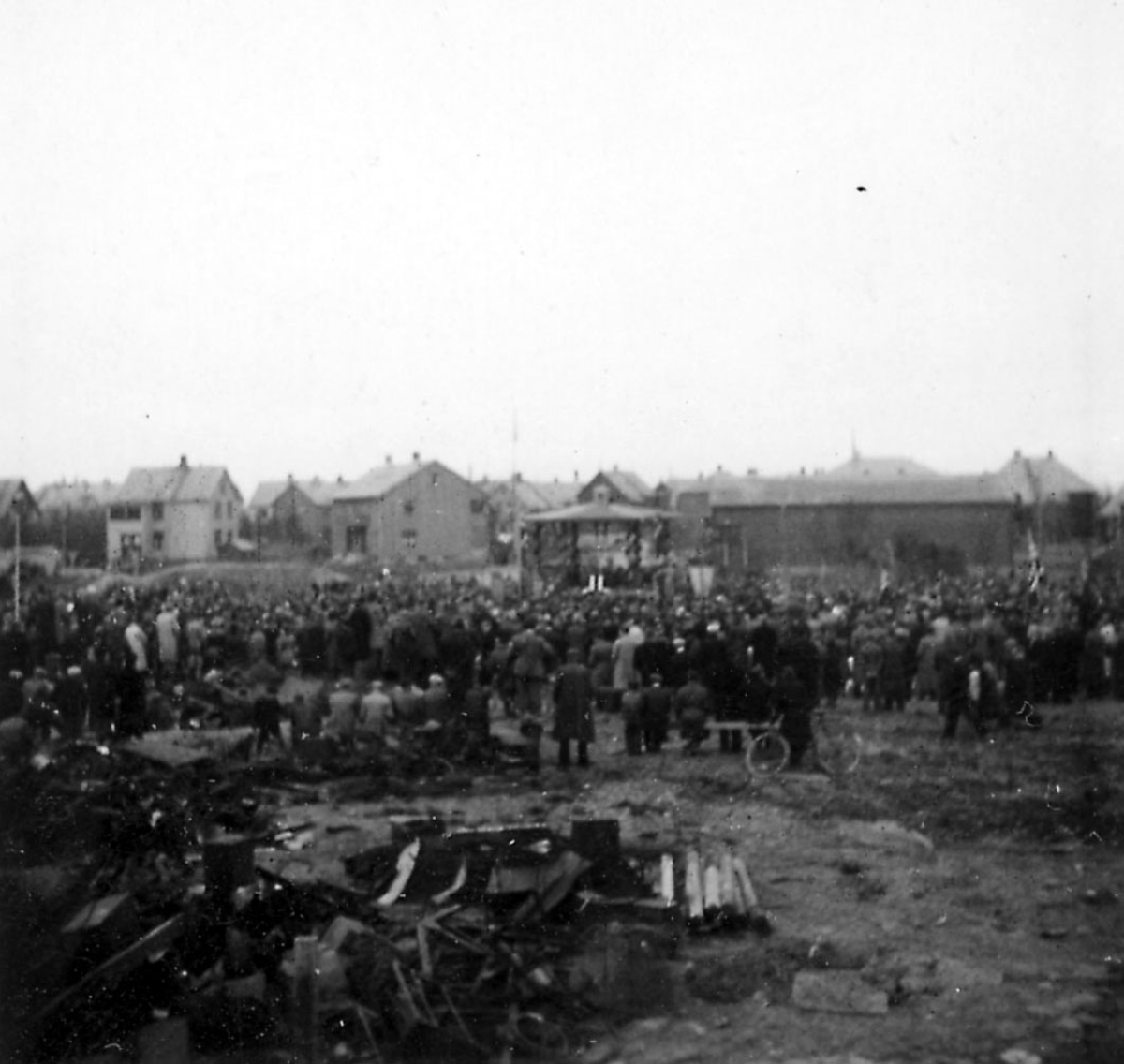 Frigjøringsdagene i Bodø etter krigen 1940 - 1945. Åpen plass med paviljong. Mange personer samlet. Rester etter ødeleggelser i forgrunnen.