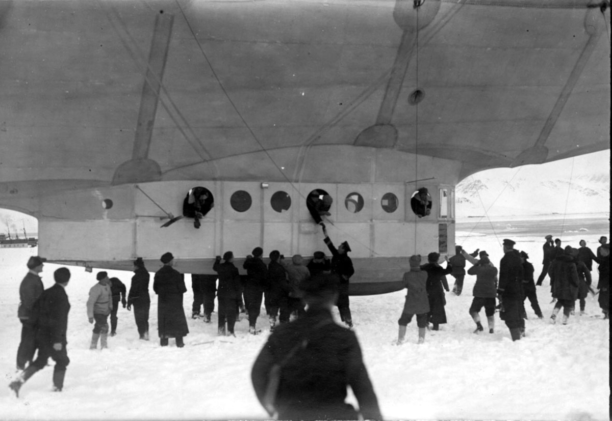 Luftskipet "Norge", undersiden med gondolen, personer i vinduene. Mange personer ved luftskipet. Fartøy ved kai, i bakgrunnen, fjorden og fjell. Snø på bakken