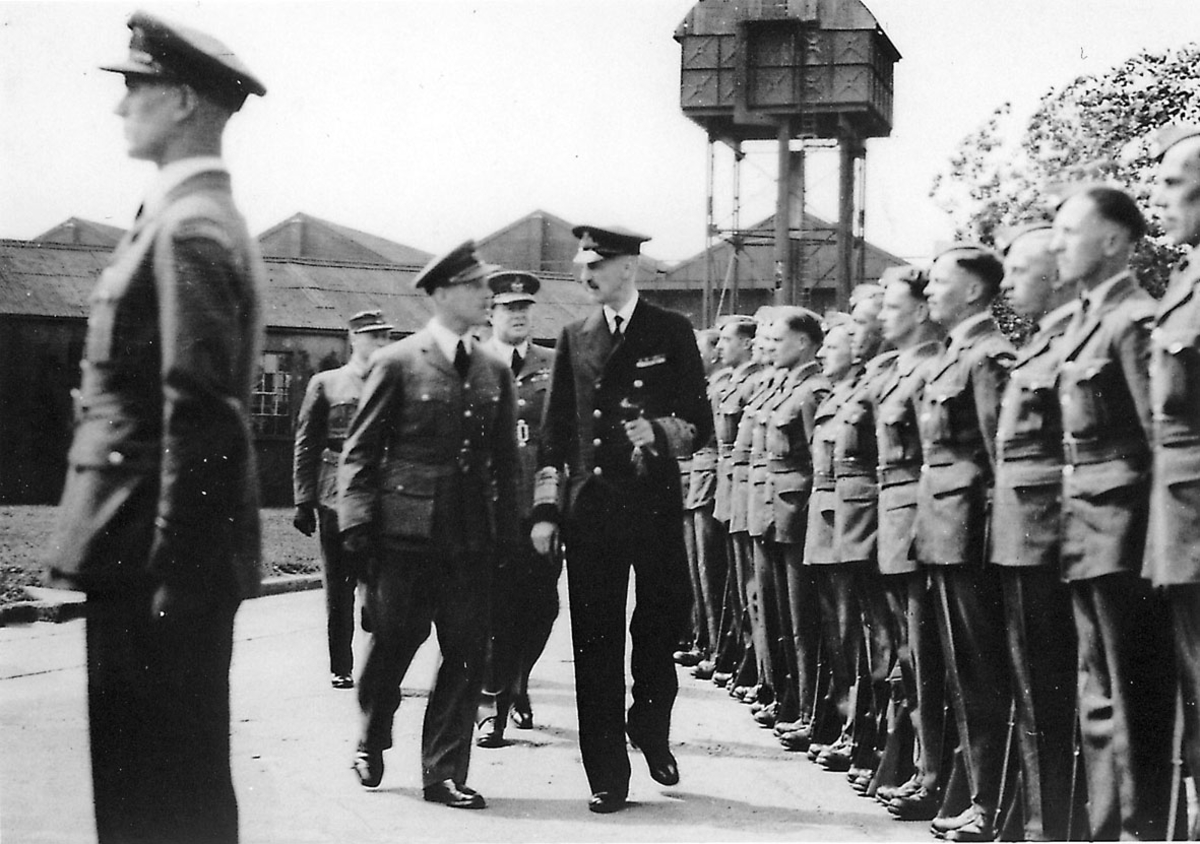 Kong Haakon VII inspiserer en gruppe soldater, fra flyvåpnet. Soldatene står oppstilt i militæruniform. Kongen i følge med uniformerte offiserer. Bygninger og "vakttårn" bak.