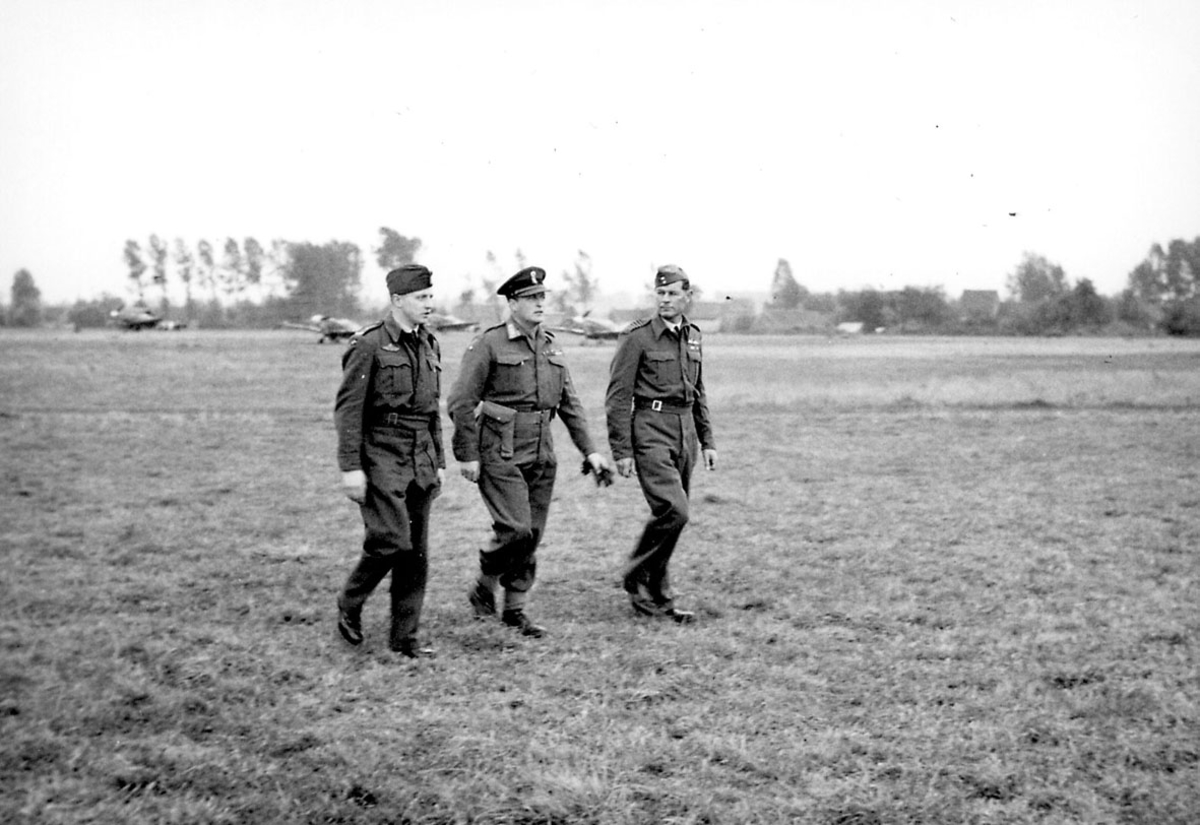 3 personer i militæruniform. Kronprins Olav sammen med 2 andre,  på en åpen plass. Noen fly i bakgrunnen.
