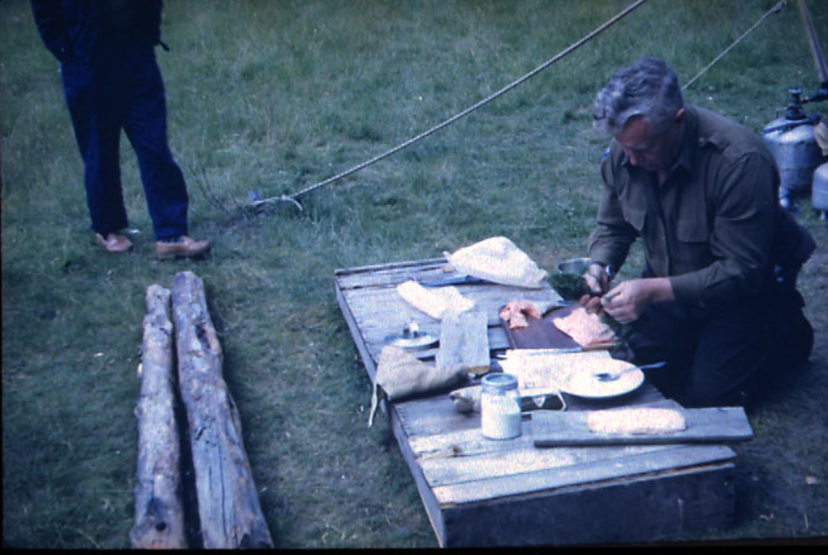 Portrett, 1 person sitter ved en benk/kasse utendørs og tilbereder fisk - laks. Føttene på 1 person sees oppe i billedkanten.