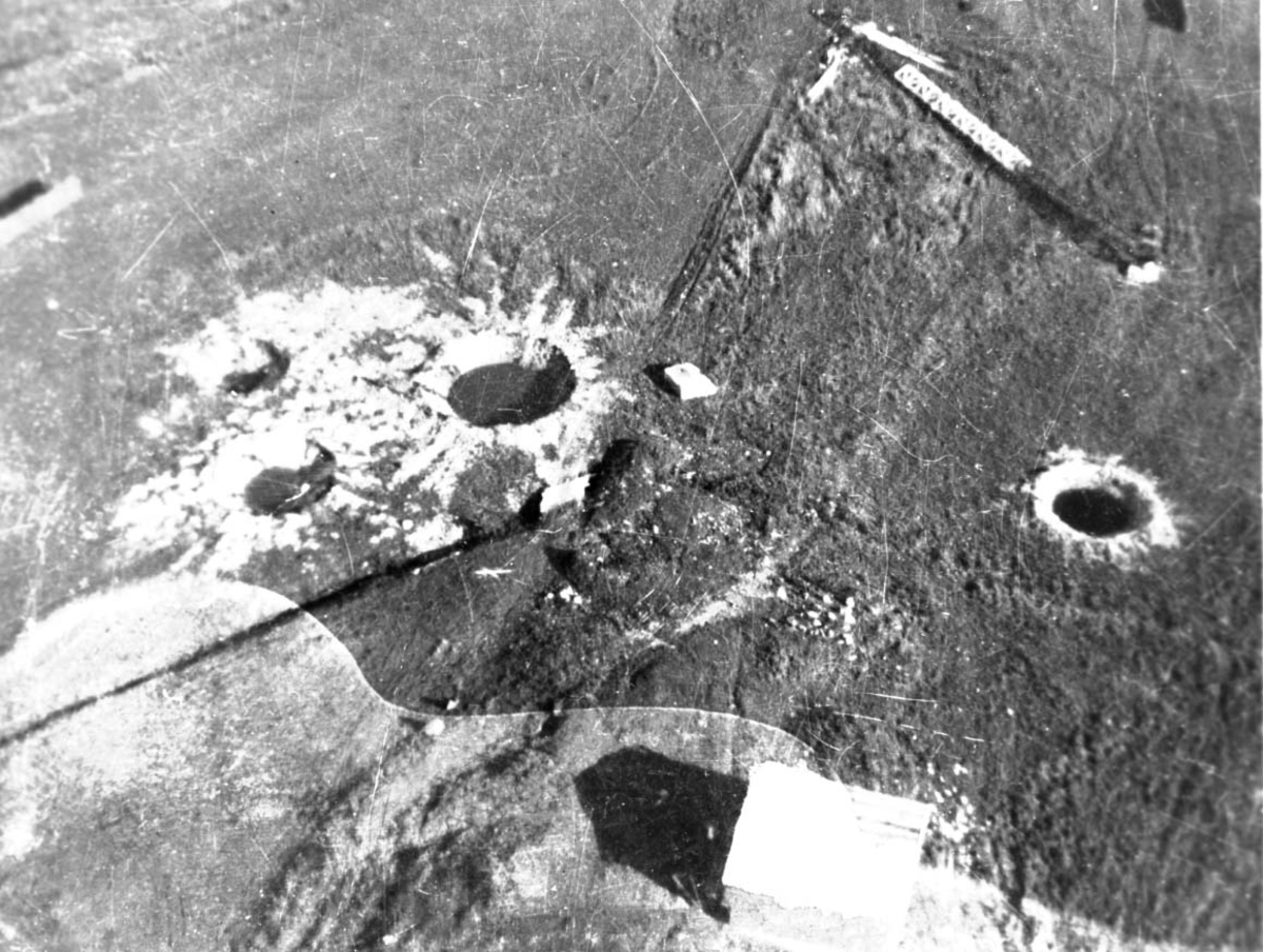 Luftfoto, flere bombekrater, store hull i bakken. Ødelagt antennemast ligger på bakken