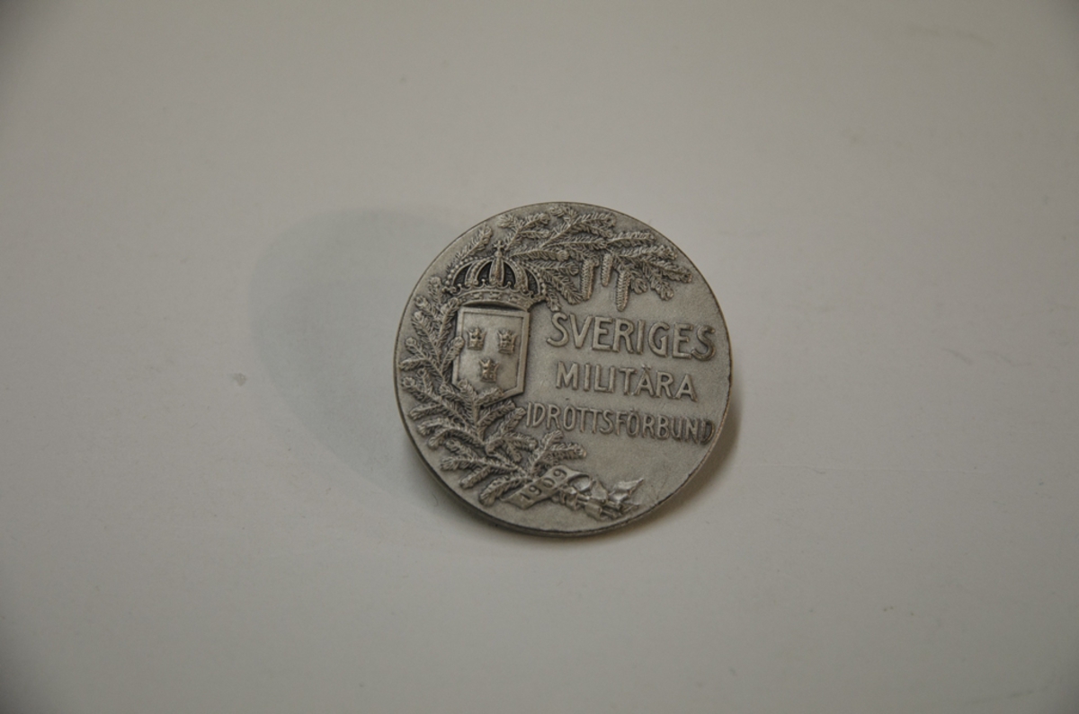 Idrottsmedalj i silver med texten: Kadettskolan YK Pistolskjutn. 9/12 1938, 1 pris. På andra sidan står det: Sveriges Militära Idrottsförbund.