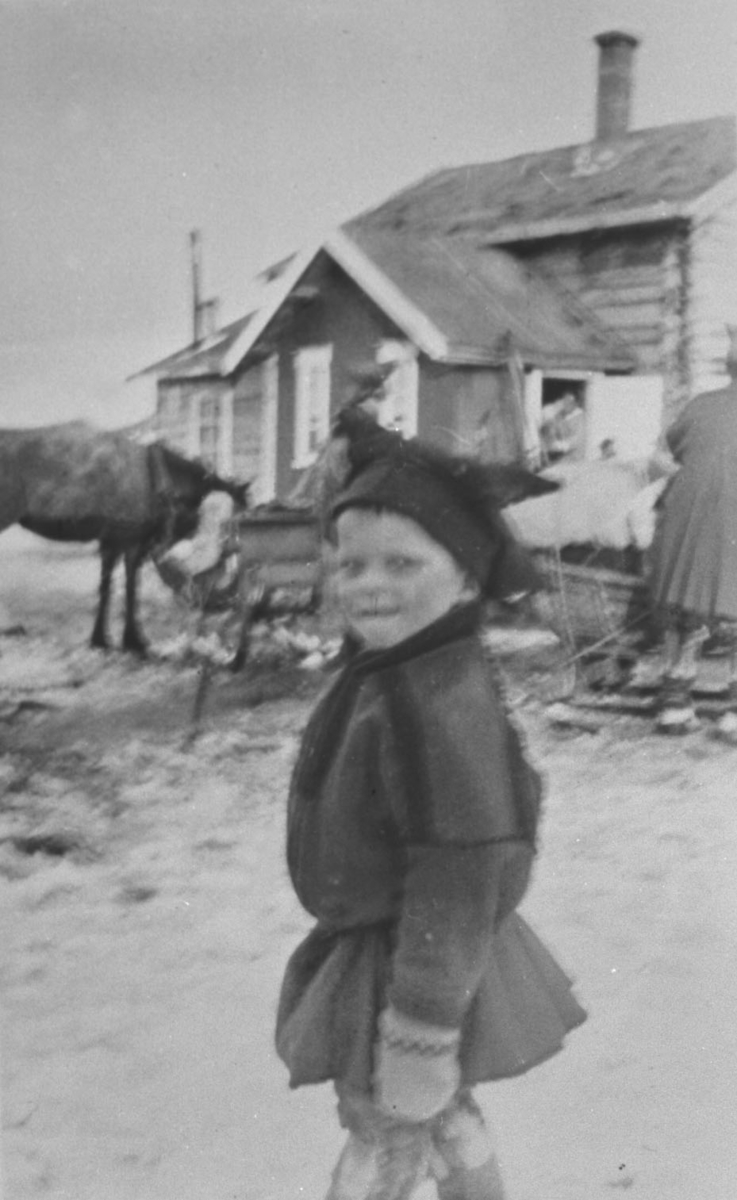Liten samegutt, Ole Olsen, står på gata trolig i Karasjok. På bakgrunn står det et hus og foran huset en hest med slede. Bildet er lite uskarp, men ellers fin.