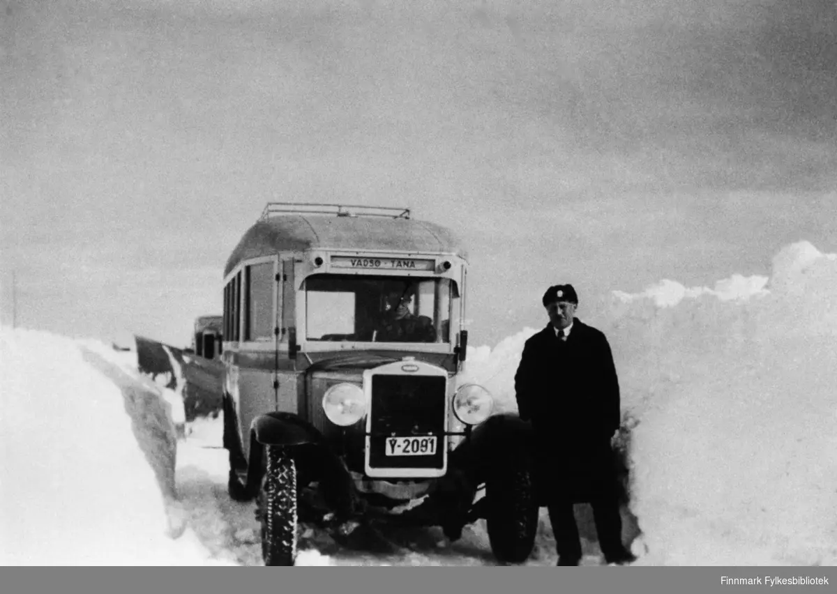 A/S Polarbil buss (y-2091, Volvo) brøyter veien, riksvei 950, Vadsø-Varangerbotn i 1940. Ved siden av bussen står en mann som heter Gustav A. Junge og en mann sitter i bussen (vi vet ikke navnet). I bakgrunnen ser man litt av brøytebilen. Se også bildene 577-588. A/S Polarbil var et rutebilselskap som eksisterte i tidsrommet 1920-1976. Ruteområdet dekte hovedstrekningene i Øst-Finnmark. Polarbil hadde hovedkontor i Vadsø. 1. januar ble selskapet med rutekonsesjoner, ansatte og bilmateriell overtatt av Finnmark Fylkesrederi og Ruteselskap (FFR).