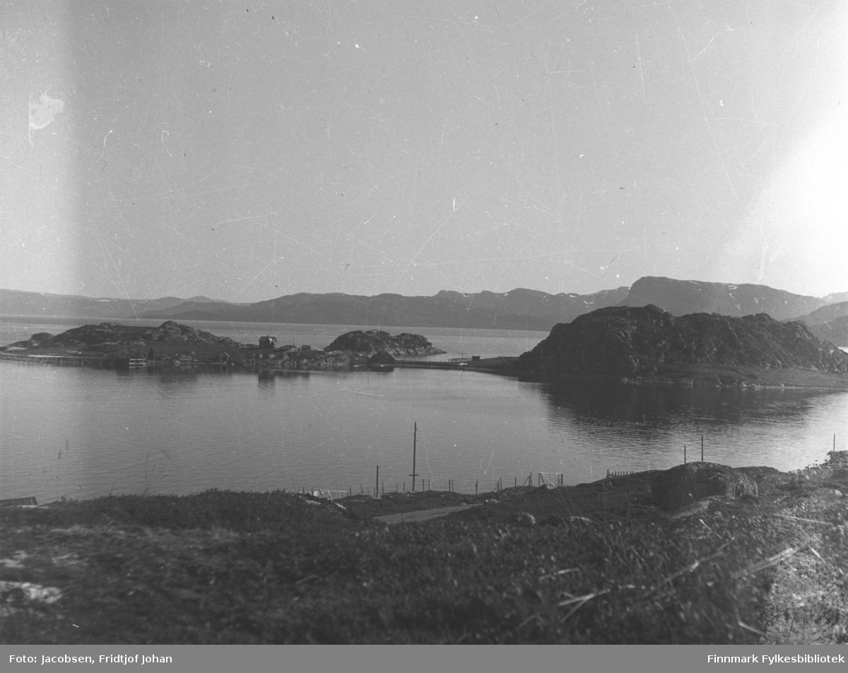 Klokkerøya, til høyre på bildet, utenfor Hammerfest/Rypefjord en sommerdag. Ved siden av Klokkerøya er Strømsnes. Bebyggelse ses langs strandkanten på halvøya. Terrenget foran på bildet er stort sett gress- og lyngdekt og Akkarfjorden har blikkstille sjø. En del stolper, både gjerdestolper og el-stolper står på området. Fjellene i bakgrunnen er Seiland. Sannsynligvis er bildet tatt før andre verdenskrig
