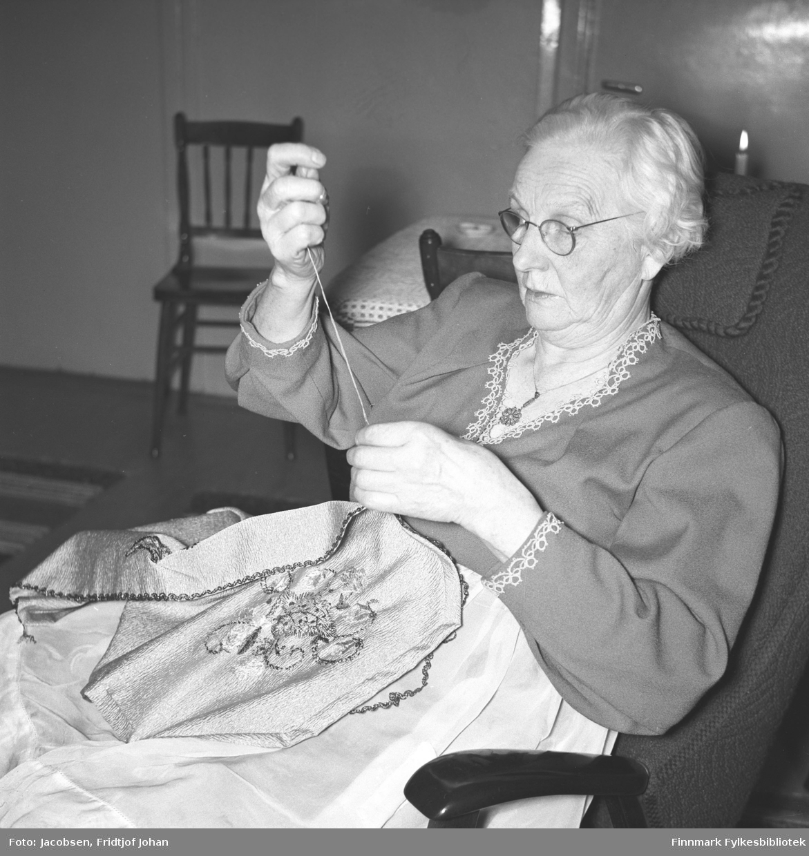 Olga Jacobsen, Arne Jacobsens farmor, sitter i en lenestol og broderer. Hun har en forholdsvis lys bluse, med mønster i halsen og rundt håndleddene, på seg. En pinnestol står ved siden av et bord bak henne. Et stearinlys brenner oppe til høyre på bildet.