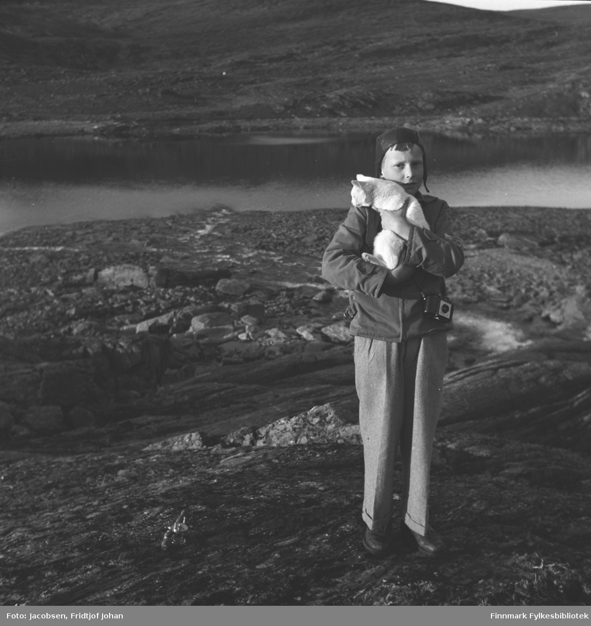 Arne Jacobsen poserer med en katt i armene. Han er iført en lys bukse, litt mørkere jakke og mørk/sort lue. Det er stort sett stein som dekker bakken der han står og et vann ses i bakgrunnen. En fjellside ses bak vannet.