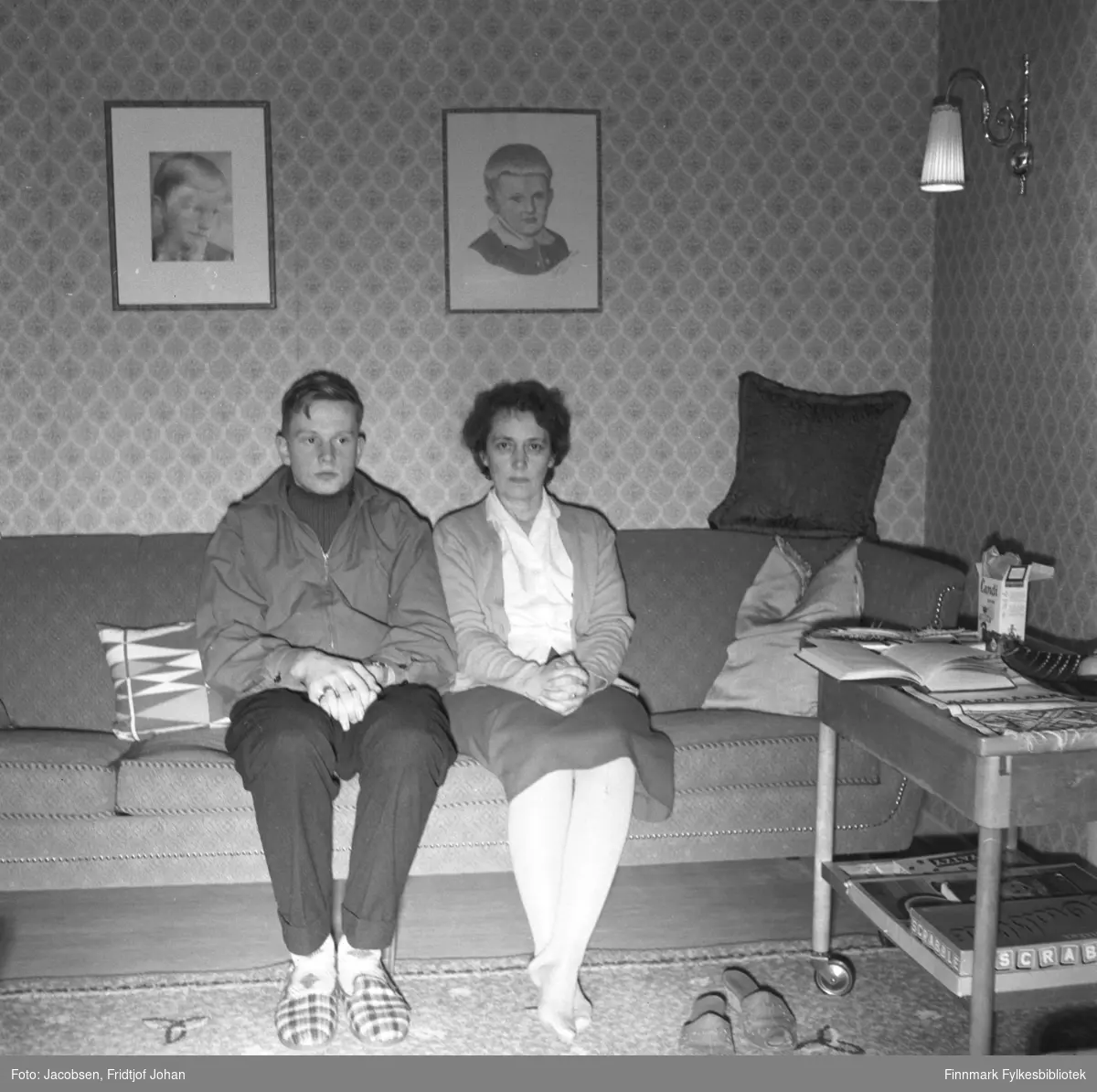Arne Jacobsen og hans mor Aase poserer for fotografen i familiens hjem i Storvannsveien. Arne har mørk bukse, genser og litt lysere jakke på seg. På føttene har han rutete tøfler. Aase har et mørkt skjørt, lys bluse og en tynn jakke på seg. Sofaen de sitter i er forholdsvis mørk og flere sofaputer ligger i den. To bilder henger på veggen bak personene og en lampe henger til høyre på bildet. Et gulvteppe ligger på gulvet foran dem til høyre står  et bord med noen spill og andre ting oppå. Tapetet er mønstret.