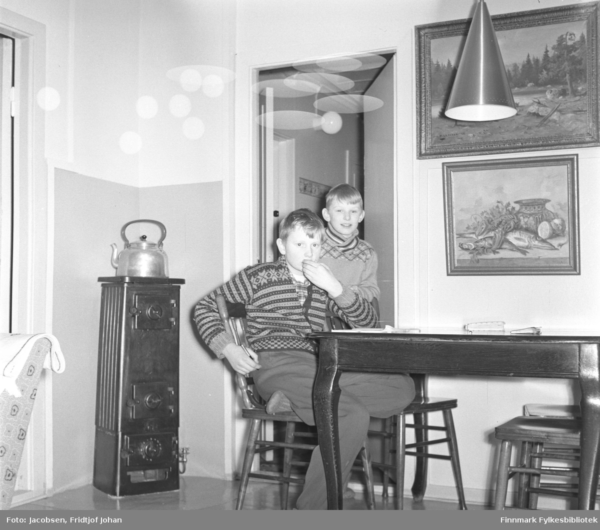 Fra stua til Fridtjof og Aase Jacobsen i Storvannsveien, vinteren 1958. På stolen sitter Arne Jacobsen. Han har en ganske lys bukse og strikkejakke med mønster på seg. Bak han står fetteren Ivar, iført en lys genser. Bordet og pinnestolene er mørke. To malerier henger på veggen over det og en taklampe ses oppe til høyre på bildet. En høy vedovn står i hjørnet med en stor kjele oppå.