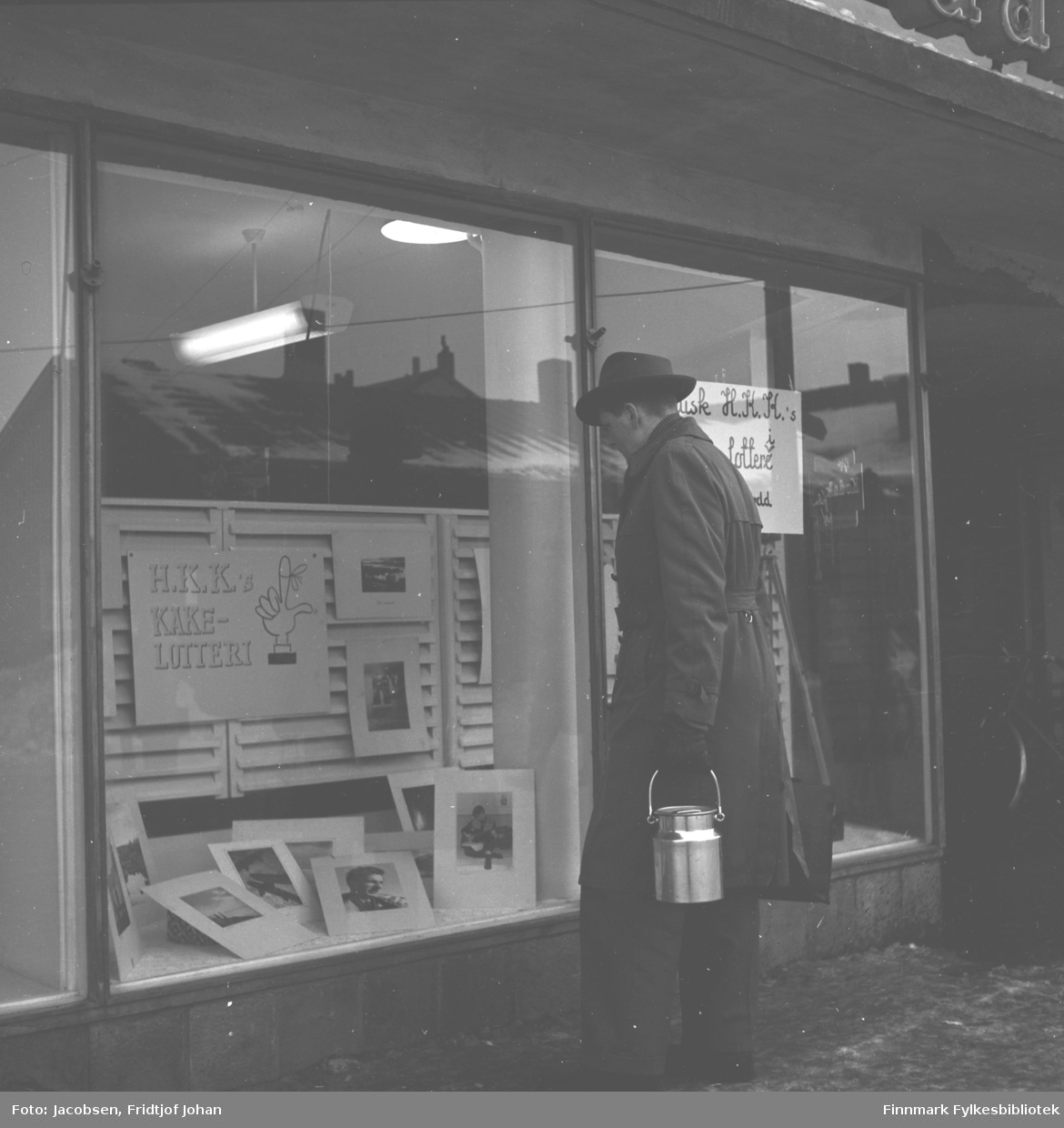 Tor Todal kikker i et utstillingsvindu til en butikk i Hammerfest. Han har mørke klær på seg, frakk/bukse og hatt. Han har en blankt spann i sin venstre hånd. Nederst i vinduet ligger flere fotografier og på veggen henger en reklameplakat for H.K.Ks kakelotteri.