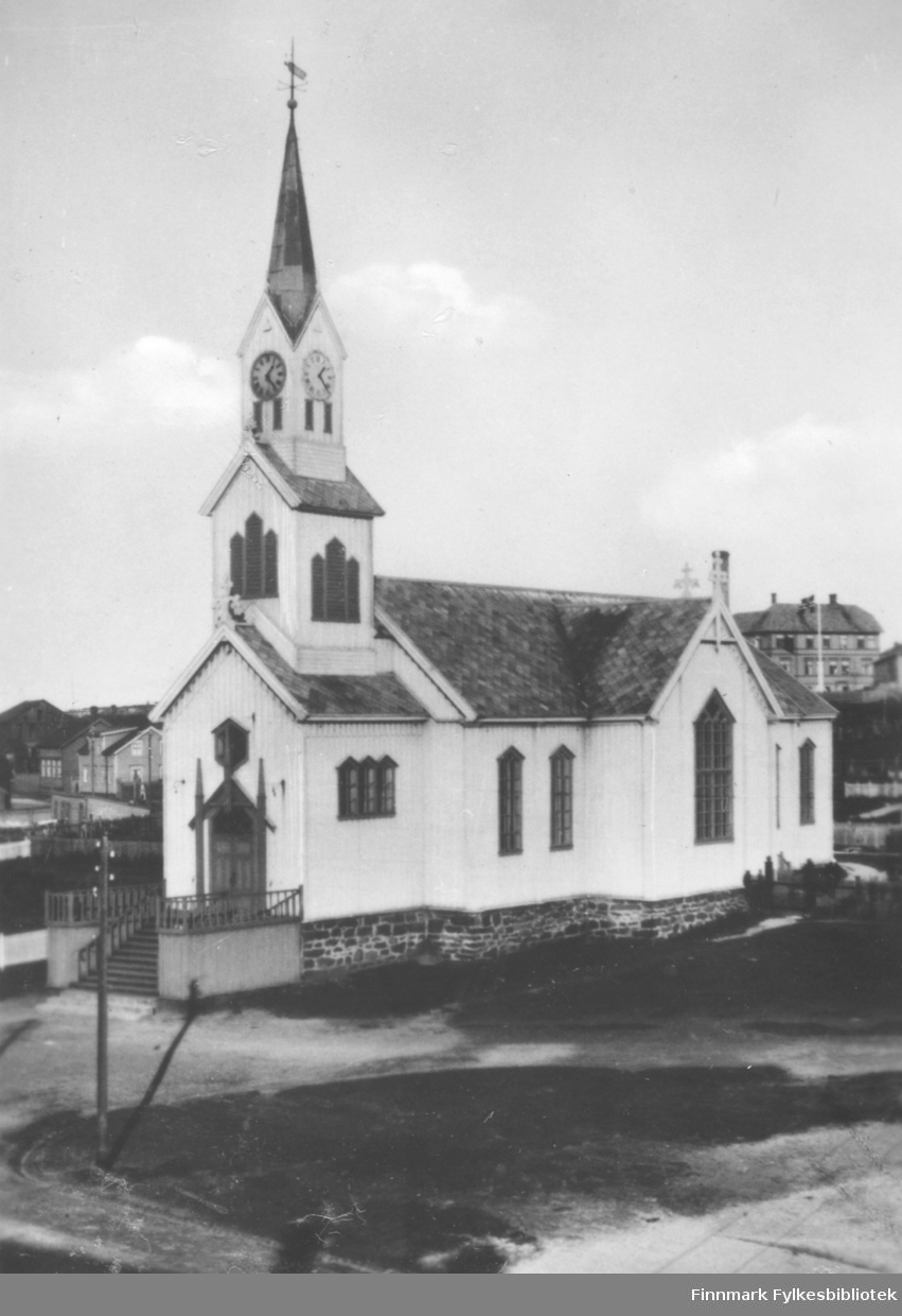 Fotografi av Vardø Kirke som var tegnet av Chr. H. Grosch og bygd i 1869. Langkirken ble brent under annen 2. verdenskrig. Til høyre bak kirken kan vi se en del av gamle Vardø sykehus, åpnet 1898. Vi ser en flaggstang foran sykehus. Eller gamle tuberkulosehjemmet?