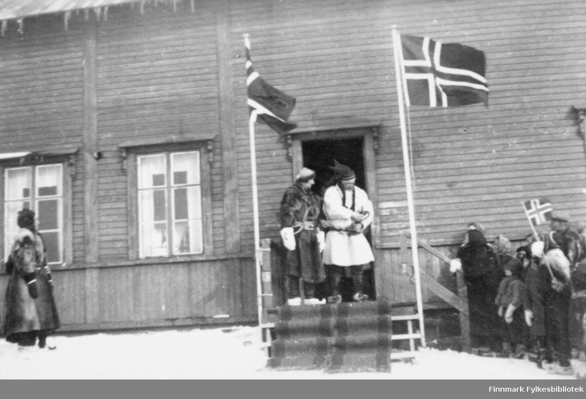 Fotografi fra kronprins Olav og kronprinsesse Martha sin reise til Finnmark i 1934. På bildet står de på en trapp utenfor en trebygning. Det norske flagg vaier oppe på to flaggstenger på hver side av trappen. To løpere ligger nedover trappetrinnene. De er kledt i skinnklær og skaller. Kronprins Olav har en samisk lue på hodet. Til venstre i bildet står det en kvinne kledt i pesk og skaller. Til høyre nedenfor en trapp står det flere mennesker og ser opp på kronprinsparret. Det er en person som holder det norske flagget. Det er snø på bakken og det henger istapper fra taket