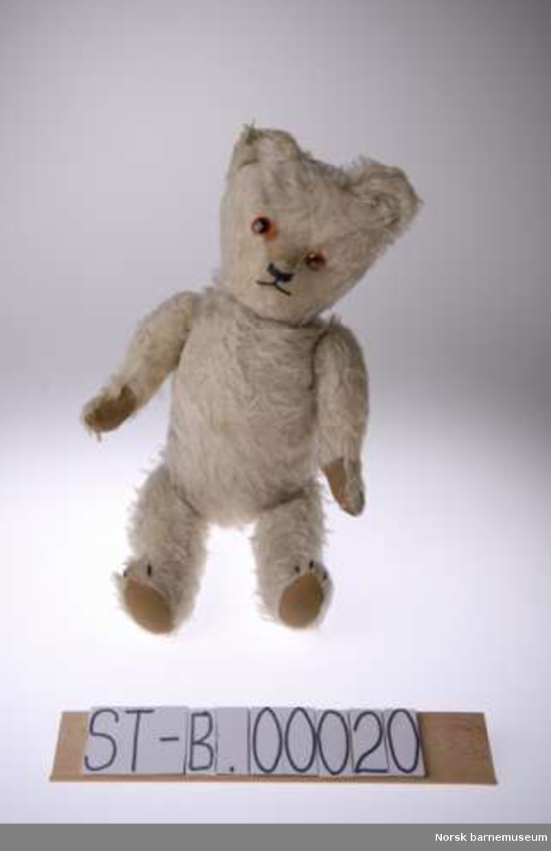 Lys teddybjørn med påsydd brunt stoff. Nese og munn markert med svart tråd. Slitt. knapper som øyne. Kan være nyere enn teddybjørnen.  