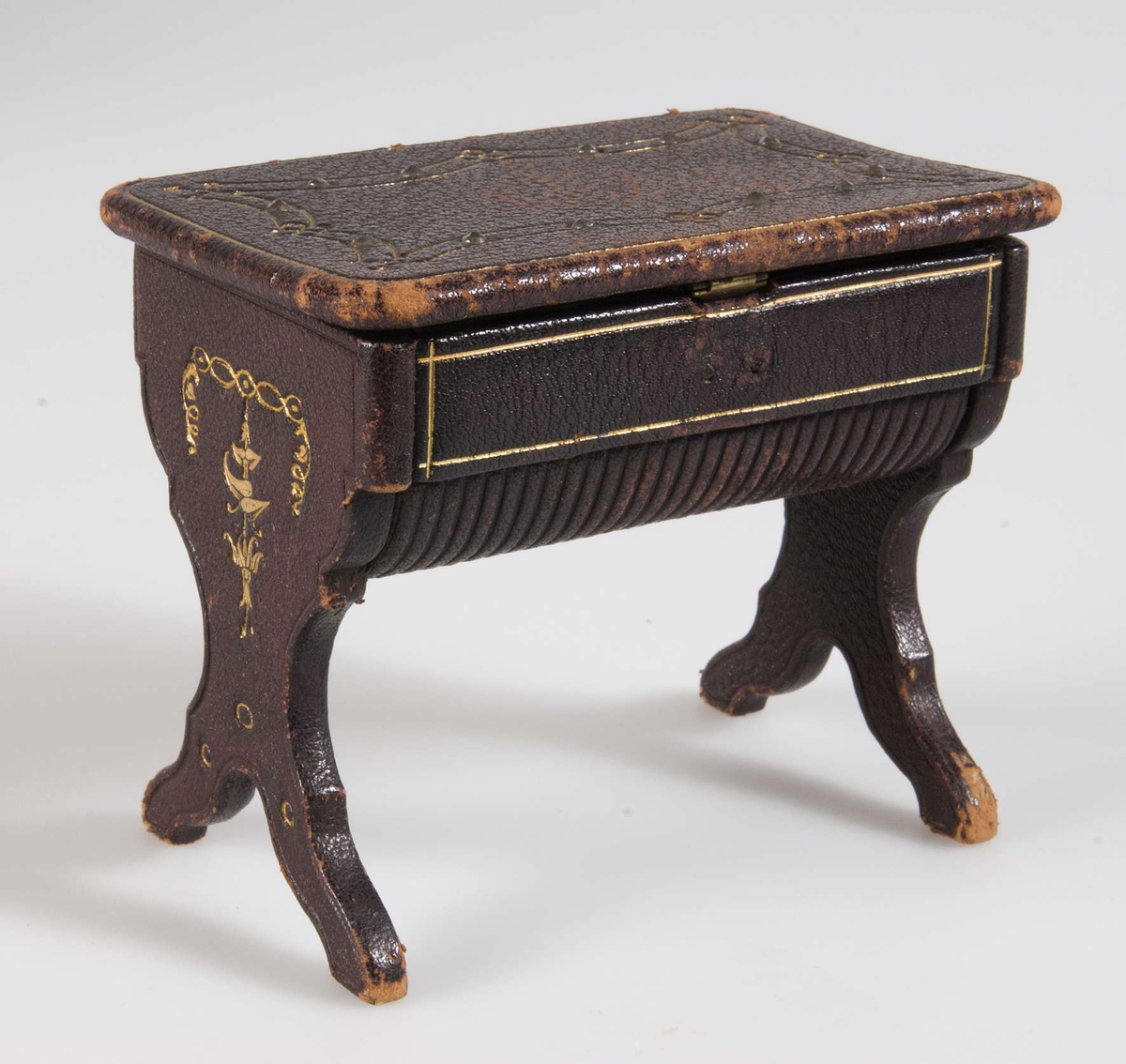 Litet sybord för dockskåp i brunt läder med innehåll: tre trådnystan, en fingerborg i metall, en bok med träpärmar, ett fodral i metall samt två vita tygbitar.