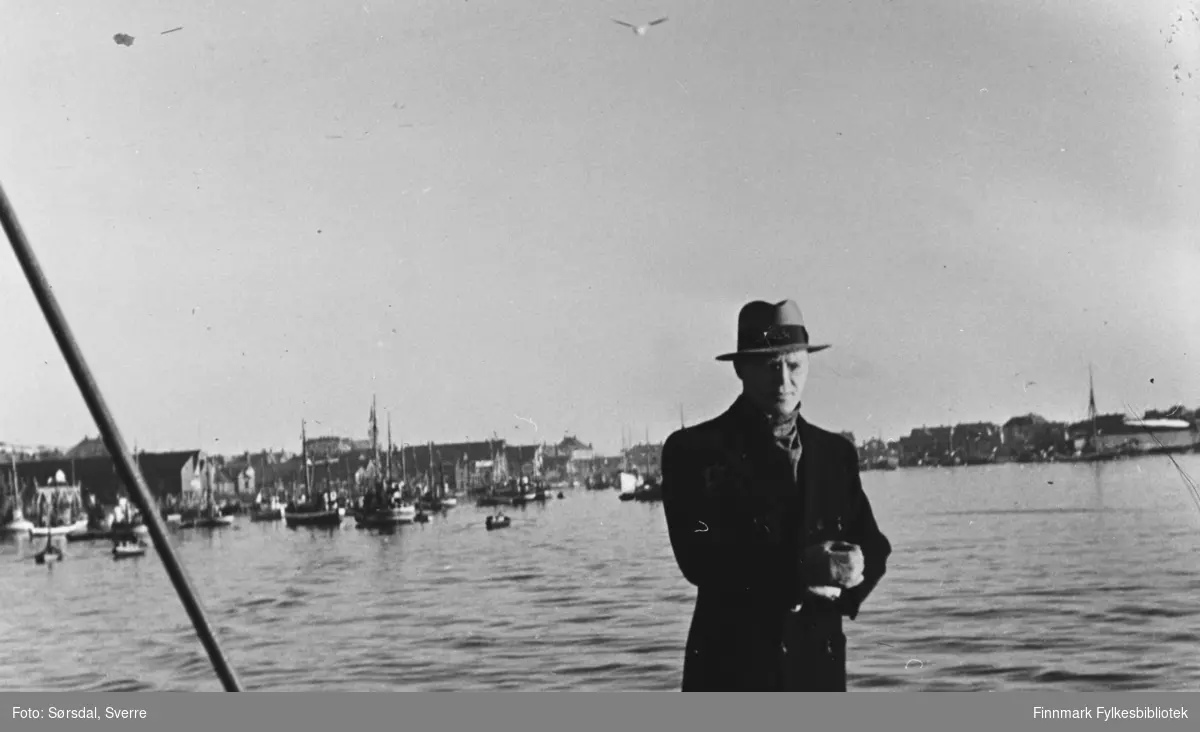Bildet over Vardø by 1934. Oversiktsbilde. Tatt fra sjøen eller moloen. I forgrunnen står en mann i frakk og hatt. Det ligger båter i havna.