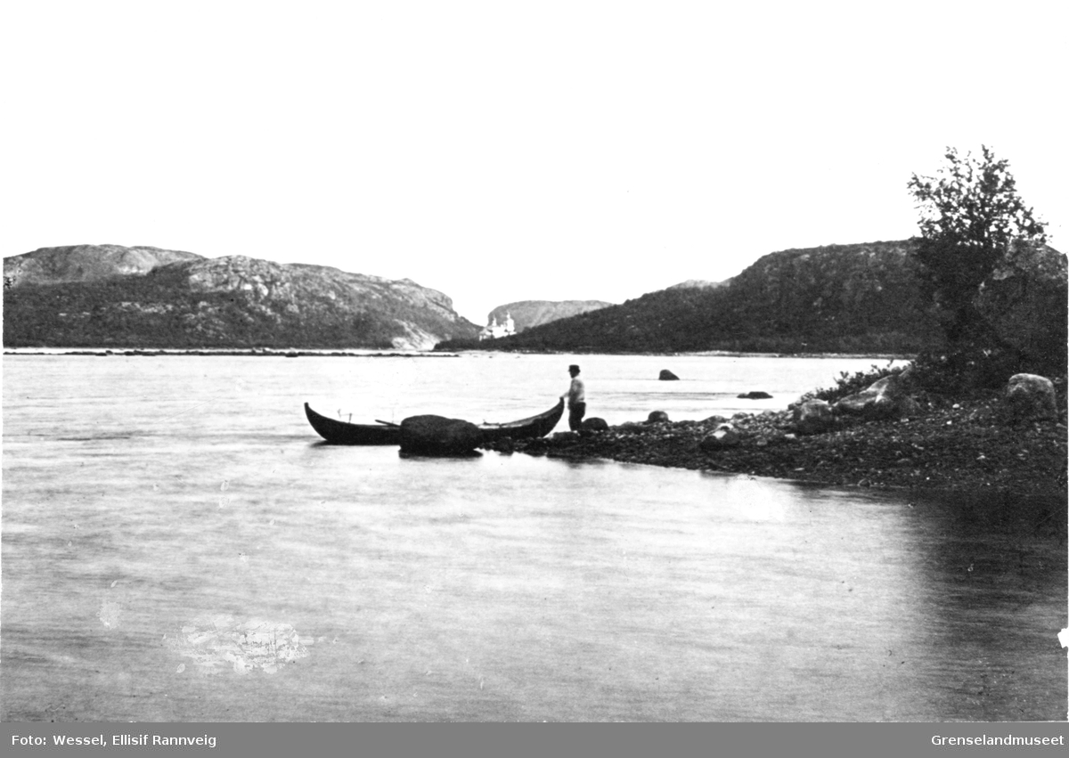 Mann og båt ved Pasvikelva nedenfor Boris Gleb, der grensen krysser elven. Kirken i Boris Gleb ses i bakgrunnen