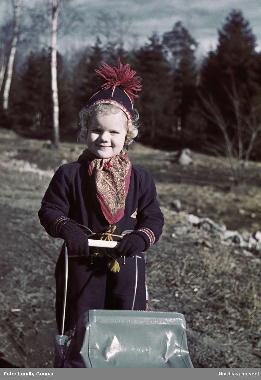 Barn i samisk-inspirerad jacka med bälte, tofsmössa och snusnäsduk i halsen, går med dockvagn ute i naturen.