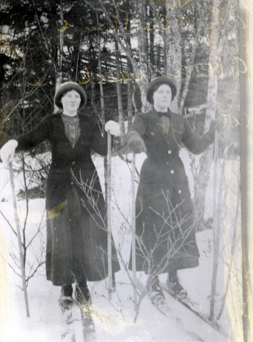 To kvinner på ski, kledd i lange kjoler (stakker) og side formsydde jakker