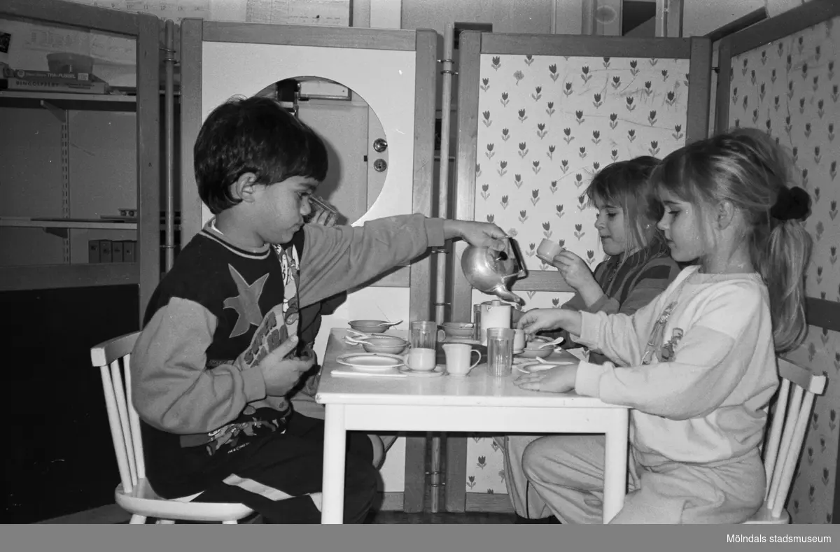 Fyra barn sitter och "fikar" vid ett barnbord. De har små kaffekoppar och fat på bordet. En pojke häller upp kaffe från en liten kittel av metall. I bakgrunden ser man en utvecklad avskärmningsdel som är klädd i olikfärgade tyger. Katrinebergs daghem, 1992.