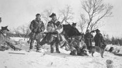 "Karasjok-samer i fullt vinterutstyr med hunder foran en lav
