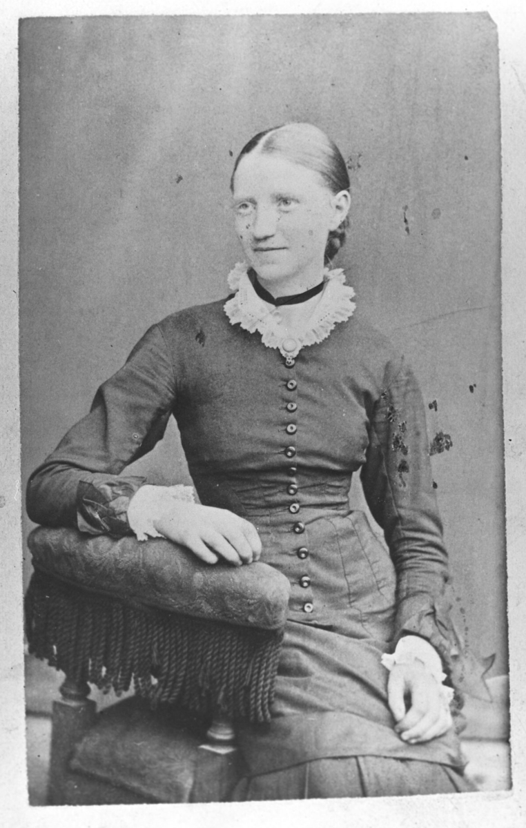 Et visitkortportrett av en ung kvinne, Tora Linseth. Hun er kledd i kjole med en lang rad av knapper foran. Hun sitter på en lenestol handen på ryggstøen og smiler pent
