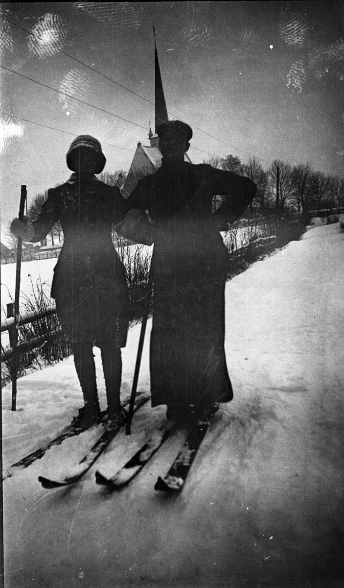 Fotoarkivet etter Gunnar Knudsen. En mann og en kvinne på skitur, kirke i bakgrunnen