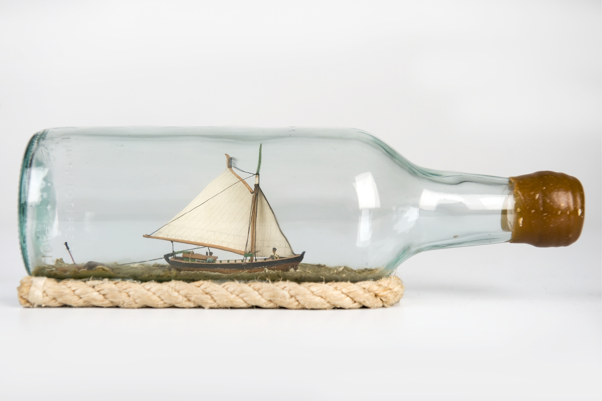Flaskskeppsmodell av sandkil under segel med jolle på släp.
Flaskan vilar på skrå av otjärat rep.