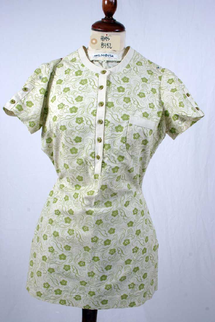 Kort kjole i hvitt bomullstekstil/bomullslerret med mønster (grønne blomster) , knappestolpe foran og påsydd liten lomme. korte ermer med knapper. 