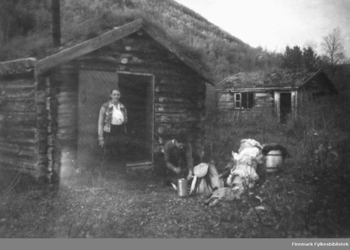 Magna Wisløff og et annet menneske foran en steinberghytte. Til høyre ser vi en lik hytte. Typiske hytter for steinbergsarbeidere i Alta. Foran ser vi reinskinn, tønne, spann og ryggsekk.