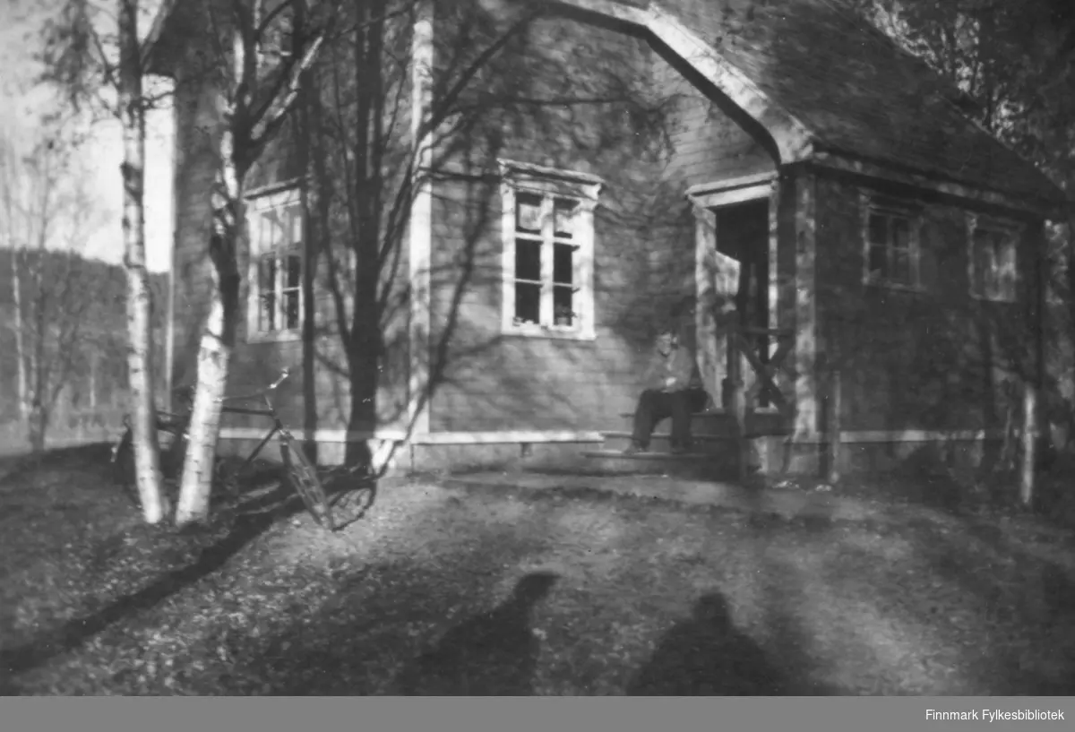 Familien Wisløffs hus før krigen. Vi ser en mann som sitter på trappa, trolig Kristian Wisløff. En sykkel står stilt opp mot noen tuntrær. Bare trær, løv på bakken og lange skygger gir bildet et høstlig preg. Ca.1930-40.